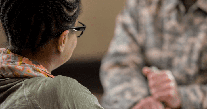 Rear view of African American female military therapist meets with male soldier. The soldier is blurred in the background. He is wearing camouflage uniform.
